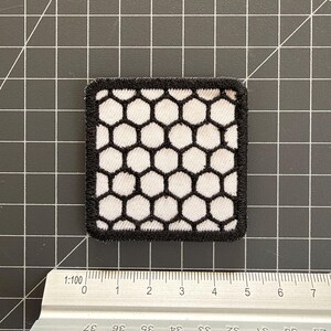May include: A square embroidered patch featuring a white honeycomb pattern outlined in black thread. The patch is positioned above a metal ruler, showing a width of approximately 1.75 inches and a height of approximately 1.75 inches.