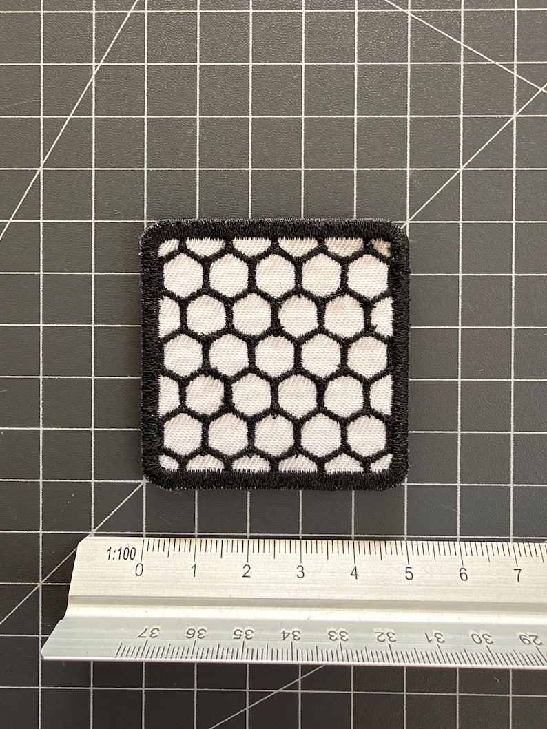 May include: A square embroidered patch featuring a white honeycomb pattern outlined in black thread. The patch is positioned above a metal ruler, showing a width of approximately 1.75 inches and a height of approximately 1.75 inches.