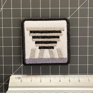 Puede incluir: Un parche bordado cuadrado con fondo blanco y un dise&ntilde;o geom&eacute;trico en negro y gris que recuerda a una estructura escalonada. El parche est&aacute; colocado sobre una regla de metal, mostrando una anchura de aproximadamente 5 cm.