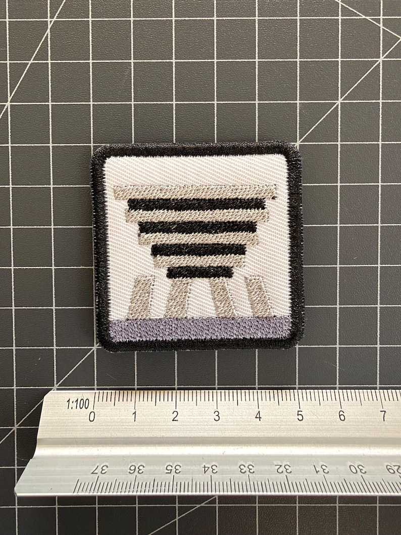 Puede incluir: Un parche bordado cuadrado con fondo blanco y un dise&ntilde;o geom&eacute;trico en negro y gris que recuerda a una estructura escalonada. El parche est&aacute; colocado sobre una regla de metal, mostrando una anchura de aproximadamente 5 cm.