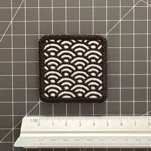 May include: A square embroidered patch featuring a black and white repeating wave pattern with a thick black stitched border. The patch is positioned above a metal ruler, showing a width of approximately 1.75 inches and a height of 2 inches.