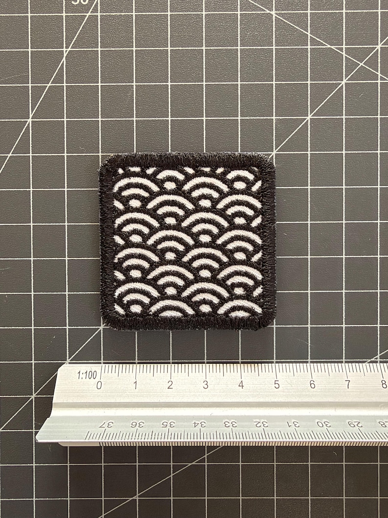May include: A square embroidered patch featuring a black and white repeating wave pattern with a thick black stitched border. The patch is positioned above a metal ruler, showing a width of approximately 1.75 inches and a height of 2 inches.