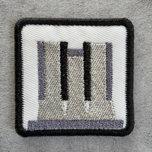 Puede incluir: Un parche bordado cuadrado con fondo blanco y un borde negro grueso. El diseño muestra una estructura industrial gris estilizada con dos columnas verticales negras prominentes y una base gris, todo ello realizado con hilo de bordar detallado.