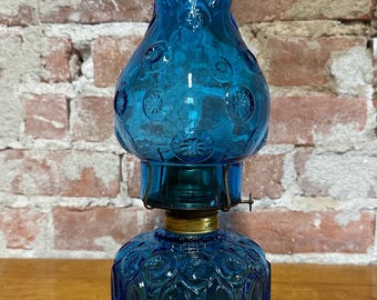May include: A blue glass oil lamp featuring a bulbous base and a matching chimney, both decorated with a repeating embossed star and oval pattern. The lamp includes a brass-colored metal burner assembly connecting the two glass sections.