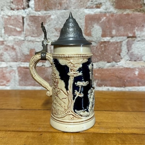 May include: A ceramic beer stein featuring a dark blue background with raised cream-colored relief designs of trees and a figure. The stein has a curved handle and a pointed, embossed metal lid with a hinged thumb lever mechanism.