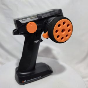 Può includere: Un radiocomando nero a impugnatura a pistola per veicoli telecomandati, dotato di un volante arancione brillante con fori circolari, una manopola di regolazione arancione e la scritta S10 DIGITAL PROPORTIONAL SYSTEM sulla base.