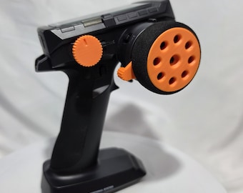 May include: A black pistol-grip radio transmitter for remote control vehicles, featuring a bright orange steering wheel with circular cutouts, an orange adjustment dial, and the text S10 DIGITAL PROPORTIONAL SYSTEM on the base.