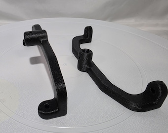 May include: Two black, textured, 3D-printed components with curved arms and mounting points. Each piece features a central cylindrical housing with a small hole and flat ends with circular openings for attachment, showing a matte finish.