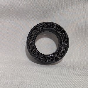 May include: A circular black ring featuring a complex, porous, and web-like internal structure. The object has a smooth outer rim and a textured, lattice-patterned surface throughout its body, creating a distinct, layered appearance.