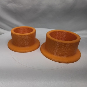 May include: Two orange cylindrical objects with a textured surface and a wide circular base. Each object features a hollow center and a smooth, flat rim at the top. The items are positioned side by side, with one being noticeably larger in diameter.