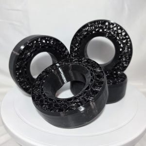 May include: Three black circular objects with a hollow center and a porous, honeycomb-like internal structure. The outer edges are smooth and solid, while the interior displays a complex, webbed pattern created through additive manufacturing.