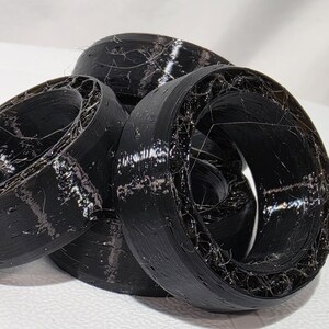 May include: A cluster of black, circular, 3D-printed rings featuring a hollow, web-like internal structure. The outer surfaces are smooth and solid, while the inner sections display a complex, fibrous, and layered filament pattern.