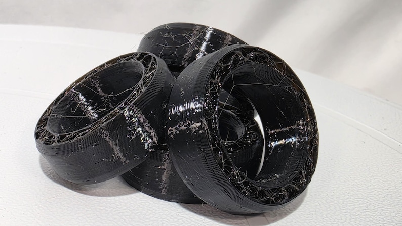 May include: A cluster of black, circular, 3D-printed rings featuring a hollow, web-like internal structure. The outer surfaces are smooth and solid, while the inner sections display a complex, fibrous, and layered filament pattern.