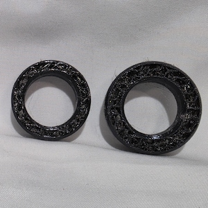 May include: Two black circular rings with a textured, porous, and layered surface finish. The rings are of different sizes, with one appearing smaller than the other. Both objects feature a distinct 3D printed appearance with visible filament strands.