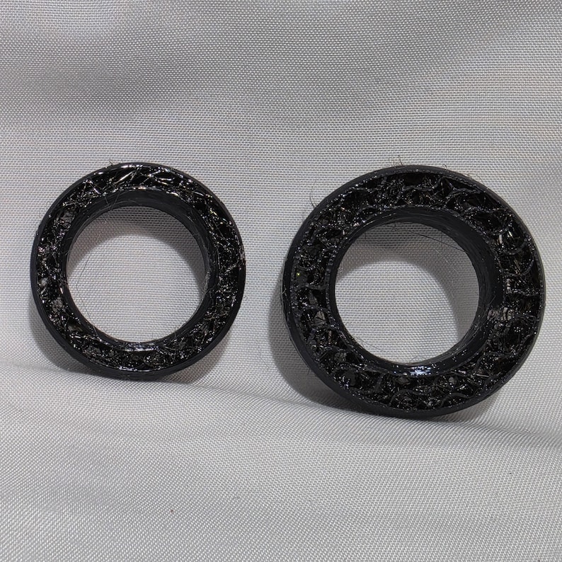 May include: Two black circular rings with a textured, porous, and layered surface finish. The rings are of different sizes, with one appearing smaller than the other. Both objects feature a distinct 3D printed appearance with visible filament strands.