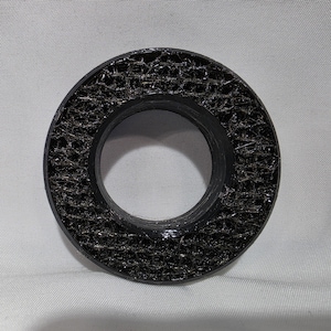 May include: A circular black object with a smooth outer rim and a textured, web-like interior pattern. The center features a hollow opening, and the surface has a glossy finish that catches the light across its intricate, mesh-like structure.