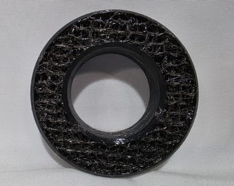 May include: A circular black object with a smooth outer rim and a textured, web-like interior pattern. The center features a hollow opening, and the surface has a glossy finish that catches the light across its intricate, mesh-like structure.