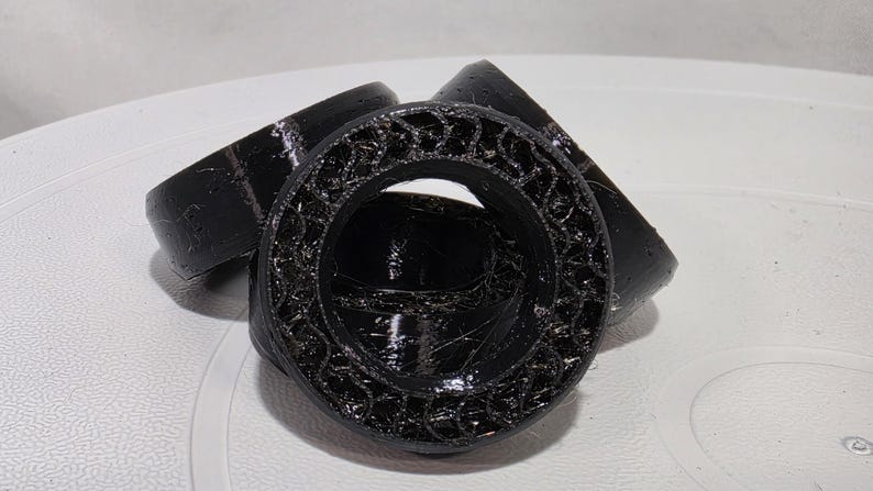 May include: Three black circular components with a hollow center and a textured, lattice-like interior structure. The outer edges are smooth and glossy, while the inner ring features a complex, repeating geometric pattern.