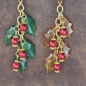 May include: A pair of dangling charms featuring gold-tone lobster clasps, gold chains, and red spherical beads with gold-tone caps. One charm displays green holly leaves, while the other features translucent amber-colored holly leaves.