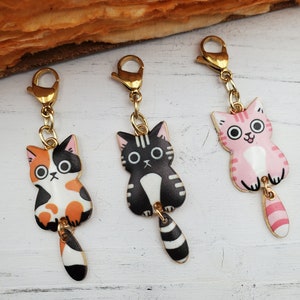 May include: Three gold charm keychains with cat designs. The cats are black and white, calico, and pink with black stripes. Each cat has a dangling tail.