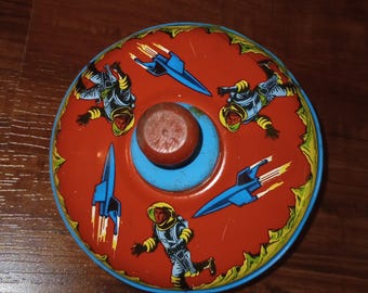 May include: A round metal spinning top toy featuring a vibrant orange surface decorated with illustrations of three astronauts in yellow and blue suits and two blue rocket ships with yellow exhaust flames. It has a central red handle.