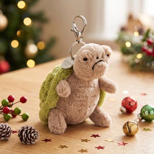 Puede incluir: Un llavero de tortuga de peluche con un cuerpo beige suave y un caparazón texturizado de color verde claro. La tortuga tiene una pequeña expresión facial triste con ojos negros y una boca curvada. Un cierre de mosquetón de metal plateado está unido.
