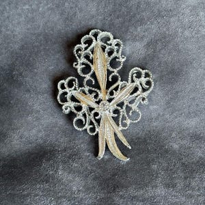Peut inclure: Une broche en métal argenté présentant un groupe central de trois formes allongées et texturées rappelant des feuilles, superposées sur un fond en filigrane ajouré complexe. Le design est symétrique avec une finition polie.