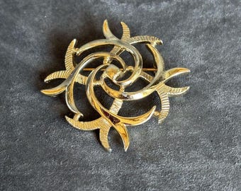 Può includere: Una spilla in metallo color oro caratterizzata da un design circolare a vortice con sei bracci curvi e appuntiti. La superficie della spilla alterna sezioni lisce e lucide ad aree testurizzate con tratteggio incrociato, creando un motivo a stella.