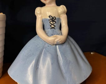 May include: A ceramic figurine of a person with brown hair, wearing a cream-coloured bodice and a wide, light blue skirt. The bodice features a black lace-up detail on the front. The figure has painted facial features, including blue eyes and red lips.