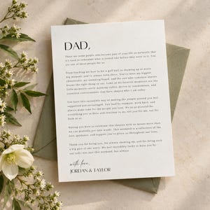 May include: A white rectangular card featuring black printed text addressed to DAD, expressing gratitude and love, signed with love by Jordan and Taylor. The card is placed on top of a sage green envelope, accompanied by white flowers.