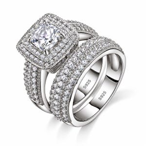 May include: A silver-toned ring set featuring a square-cut center stone surrounded by a double halo of small clear stones. The bands are encrusted with multiple rows of pavé-set clear stones and are clearly marked with the S925 hallmark inside.