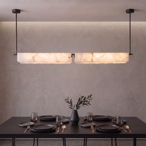 May include: A linear pendant light fixture featuring two rectangular, translucent white stone panels suspended by thin black metal rods. The fixture emits a warm glow above a black dining table set with dark plates, glasses, and gold cutlery.