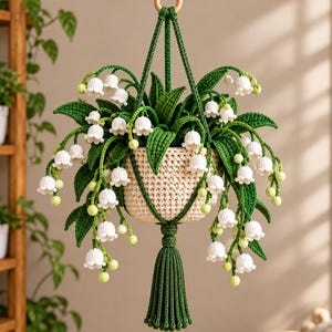 May include: A crocheted hanging plant featuring white lily of the valley flowers and green leaves in a cream-coloured pot. The plant is suspended by green crocheted cords attached to a wooden ring at the top and finished with a green tassel.