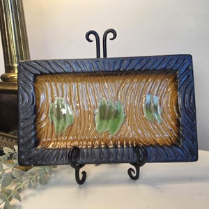 May include: A rectangular ceramic tray featuring a dark brown textured border and a tan center with vertical grooves. Three distinct green glazed patches are centered horizontally across the tray, which is displayed on a black metal stand.