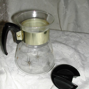 May include: A vintage glass coffee carafe with a black handle and lid. The carafe has a gold band around the middle and is decorated with starburst designs. The carafe is sitting on a white floral patterned surface.