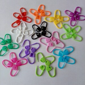 May include: A collection of colorful plastic stitch markers in various shades, including red, orange, yellow, green, blue, purple, pink, white, and black. The markers are in an open, clasp-like design, ideal for knitting and crochet projects.