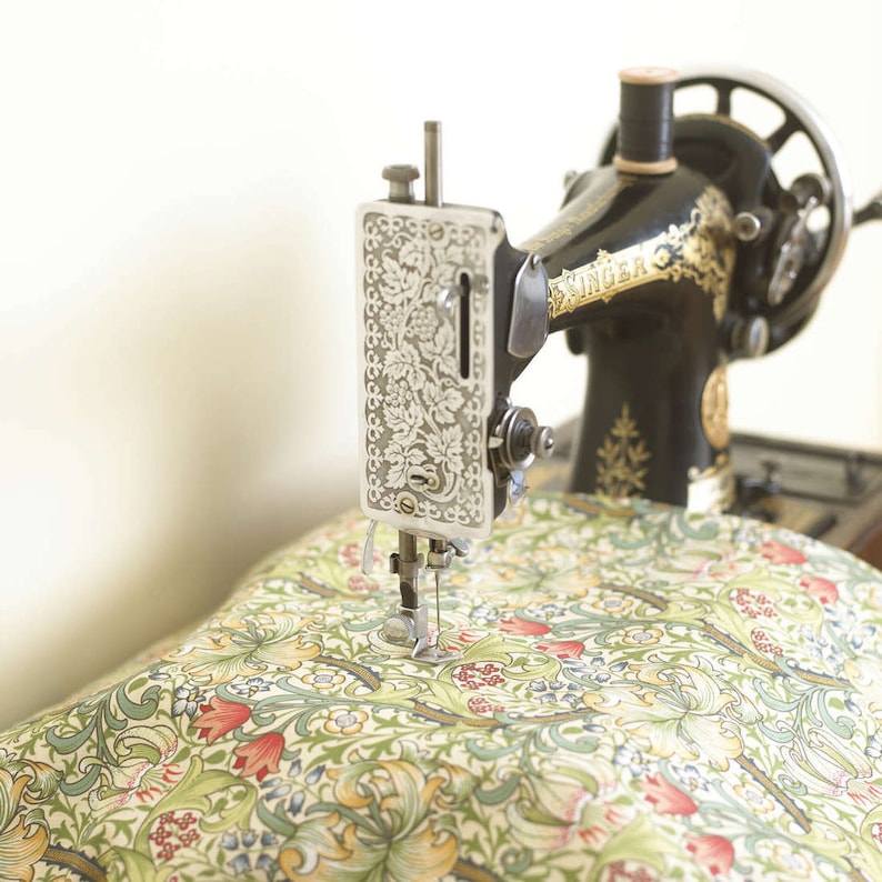 Puede incluir: Una m&aacute;quina de coser Singer vintage negra con detalles dorados est&aacute; cosiendo una tela estampada con flores rojas, verdes y amarillas. La m&aacute;quina tiene una aguja plateada y una bobina de hilo.