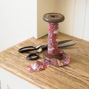 May include: A decorative wooden spool wrapped in red patterned fabric, alongside a pair of black scissors. The spool and scissors are arranged on a wooden surface, with some fabric unwound.