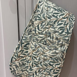 May include: A double oven glove featuring a botanical leaf design in green and brown on a cream background. The quilted glove has a hanging loop and is designed for kitchen use.