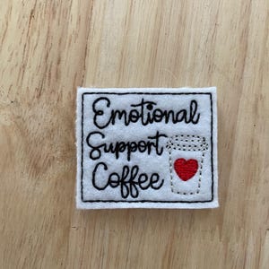 May include: White felt patch with black embroidered text that reads "Emotional Support Coffee" and a black embroidered outline of a coffee cup with a red heart inside.