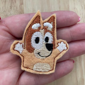 May include: A brown and white felt finger puppet of a cartoon dog with big eyes and a smile. The dog is wearing a brown collar.