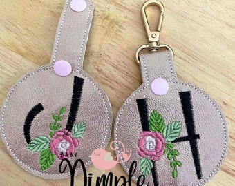 May include: Two gold-colored keychains with a floral design and a letter embroidered on each. The keychain on the left has the letter "J" and the keychain on the right has the letter "H". Both keychains have a pink flower and green leaves. The text "Baby" is embroidered on the keychain on the left. The text "Dimple Designs" is printed below the keychains.