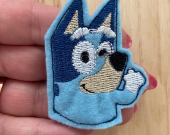May include: Blue and white embroidered felt patch of a cartoon dog with a black eye patch. The dog is smiling and has a white patch on its forehead.