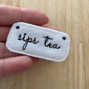 Puede incluir: Parche de fieltro blanco con bordado negro que dice "Sips Tea" con una estrella a cada lado.