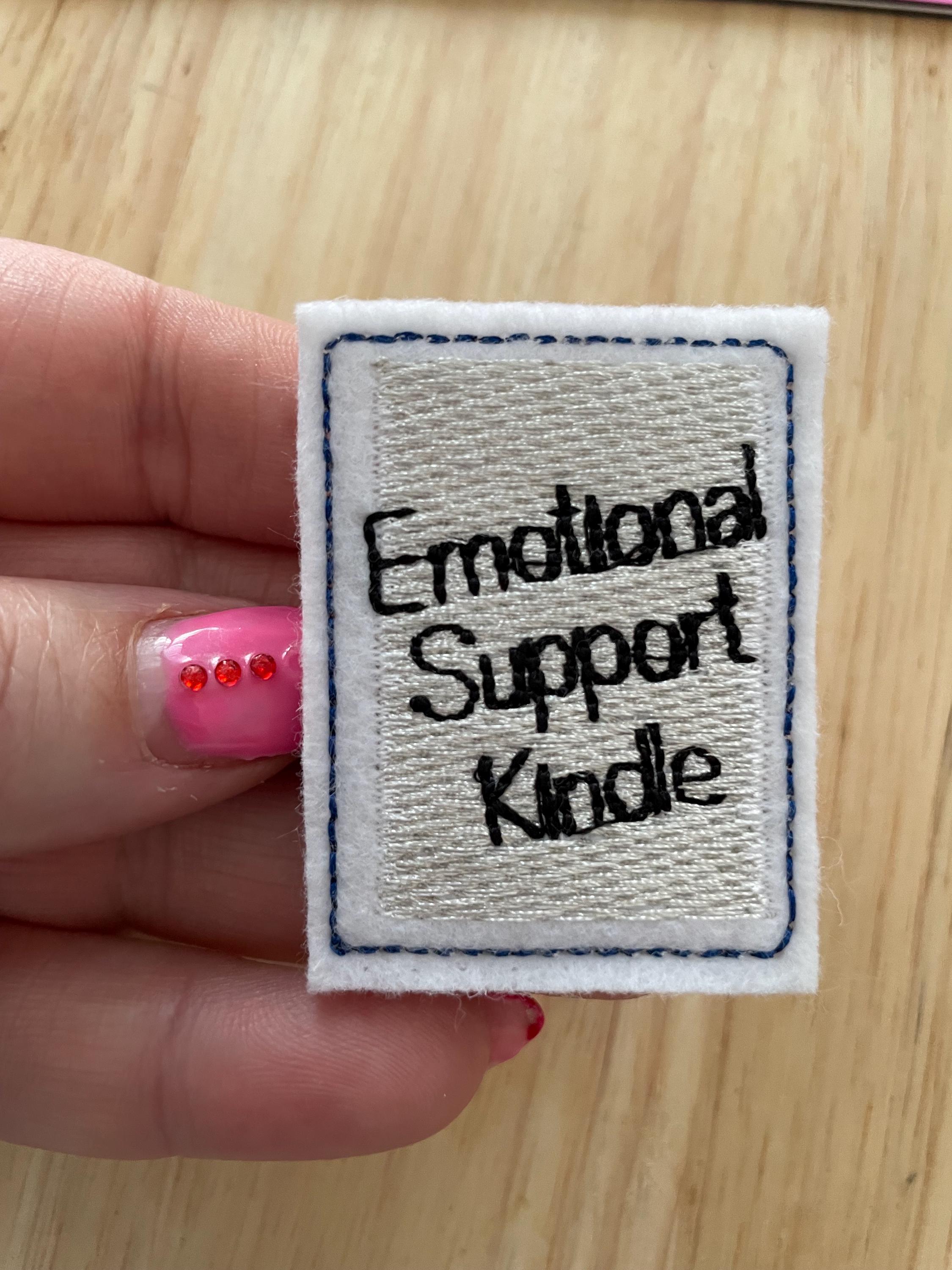 May include: A rectangular, embroidered patch with the words "Emotional Support Kindle" in black thread on a white background. The patch has a blue border and is made of felt.
