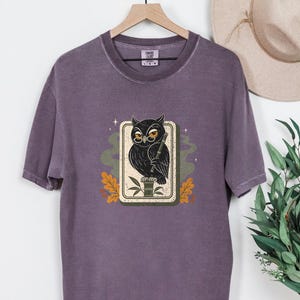 May include: A dark purple short-sleeve t-shirt featuring a graphic print of a black owl wearing round glasses and holding a bamboo stalk. The owl is centered within a rectangular frame, surrounded by stylized green smoke, small stars, and orange leaves.
