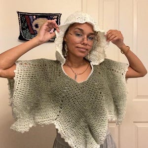 May include: A crochet hooded poncho featuring an ombre color transition from a deep sage green at the center to a creamy off-white at the edges. The garment has a V-neckline, short wide sleeves, and a scalloped hemline with a delicate openwork pattern.