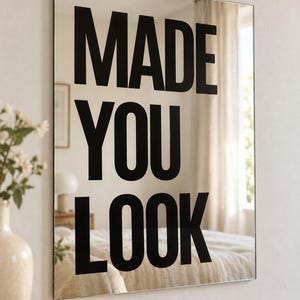 May include: A rectangular wall mirror with a reflective surface featuring the bold black text MADE YOU LOOK printed in large capital letters. The mirror has a thin, clean edge and is mounted on a wall, displaying a clear reflection.