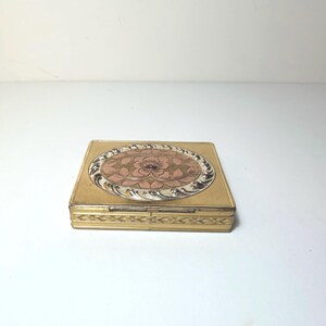May include: A rectangular gold-toned metal compact case featuring a central oval inlay with a pink floral design surrounded by a silver-toned twisted rope border. The sides of the compact are decorated with a repeating embossed leaf pattern.
