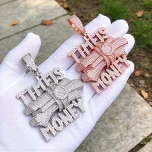Puede incluir: Dos colgantes de metal con forma de fajo de billetes atado con una banda, con el texto "TIME IS MONEY" en piedras engastadas en pavé. Un colgante es de tono plateado y el otro de oro rosa, ambos cubiertos de pequeñas piedras blancas brillantes.